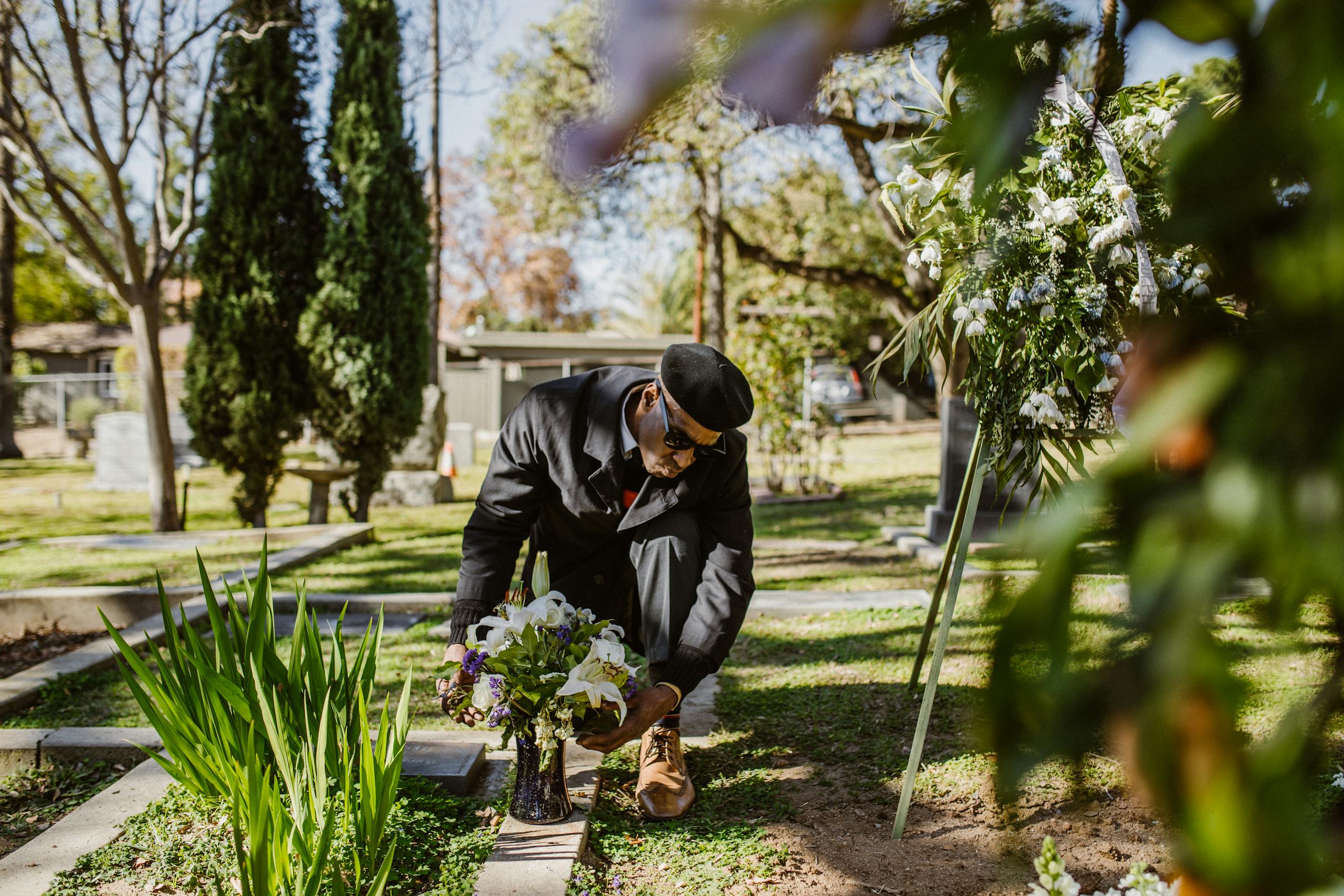 A man in black clothing mourns by a gravestone in a sunlit cemetery, surrounded by greenery and flowers.